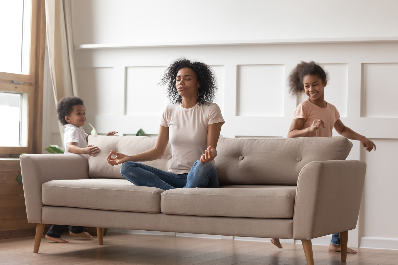 Calm african mom doing yoga on sofa relaxing with kids
